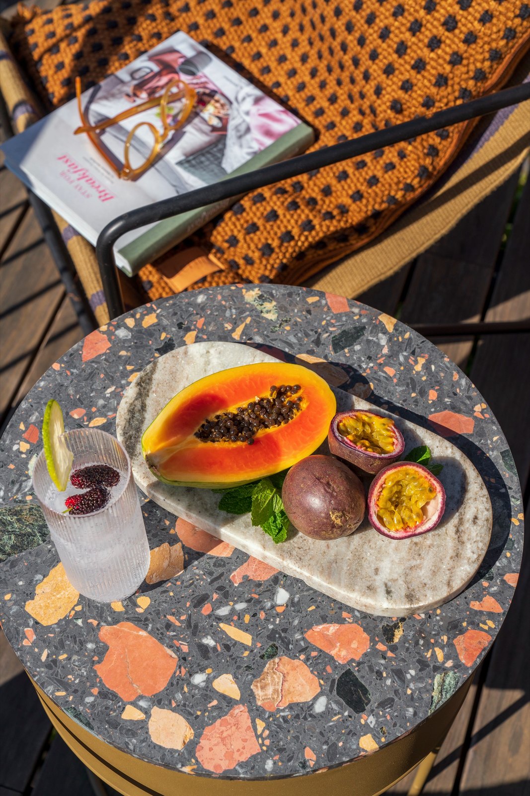 Botânica Living — Terrazzo side table with a stone board of tropical fruit and a drink, beside a woven chair