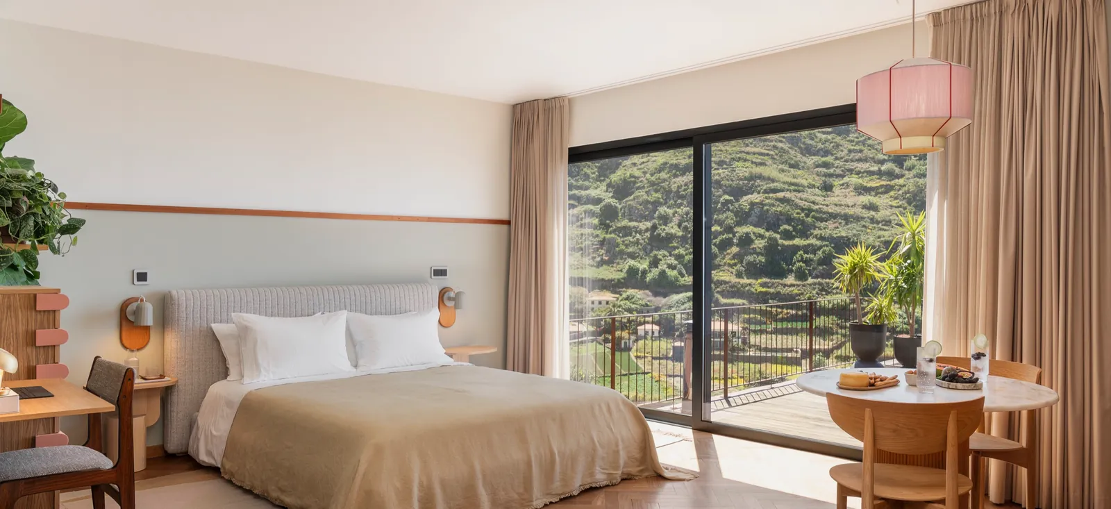 A bedroom at Botânica Living: a bed with a linen throw, a pink pendant light over a round table, and a glass wall opening onto a balcony and terraced green hillside