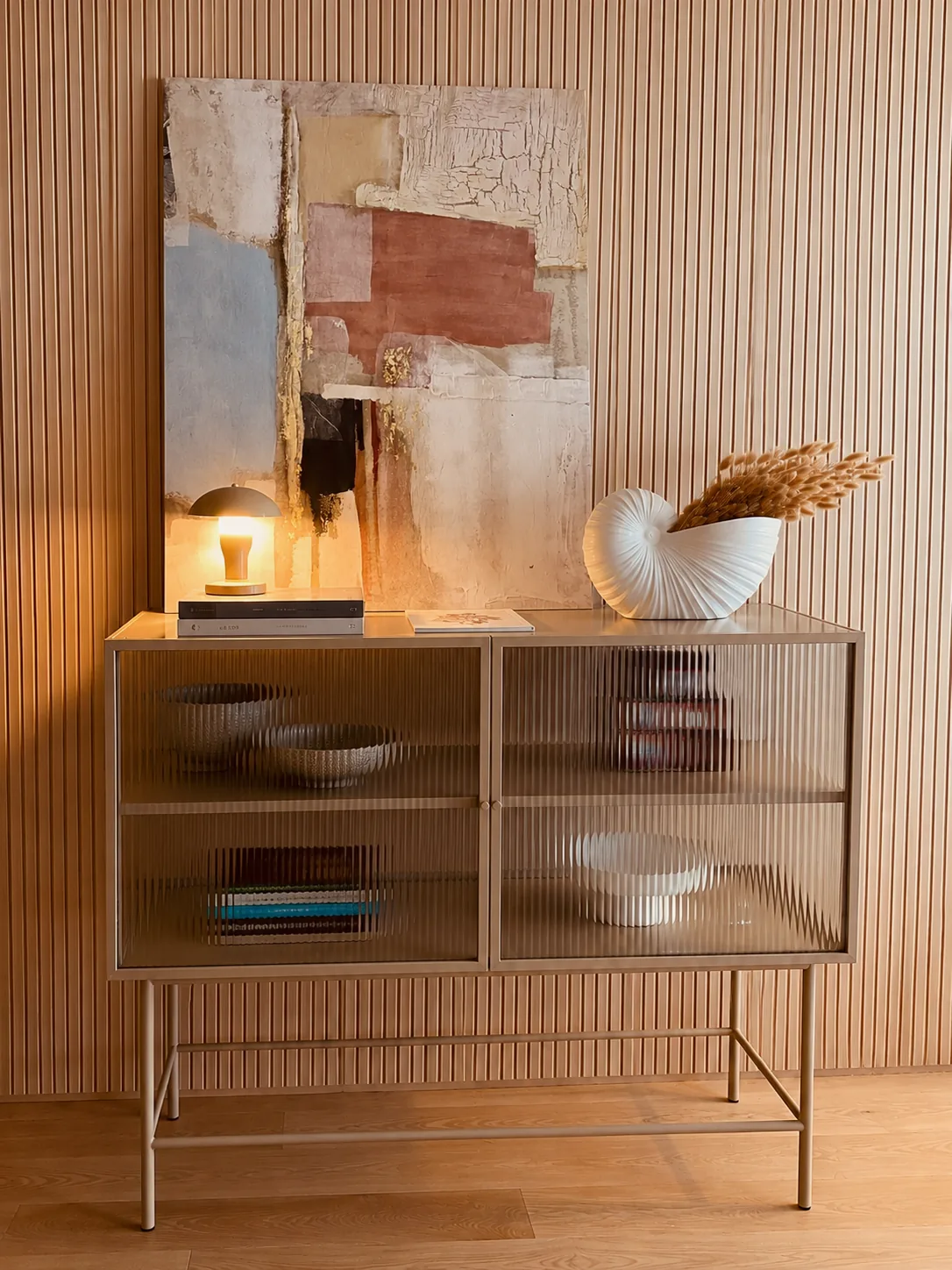 Private Residence — a fluted-glass sideboard against oak slats, with an abstract painting, a small lamp and a white shell vase with dried grasses