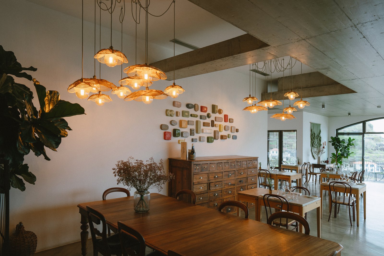 Socalco Nature Calheta — Dining room with clusters of woven pendant lamps, wooden tables and a wall of ceramics