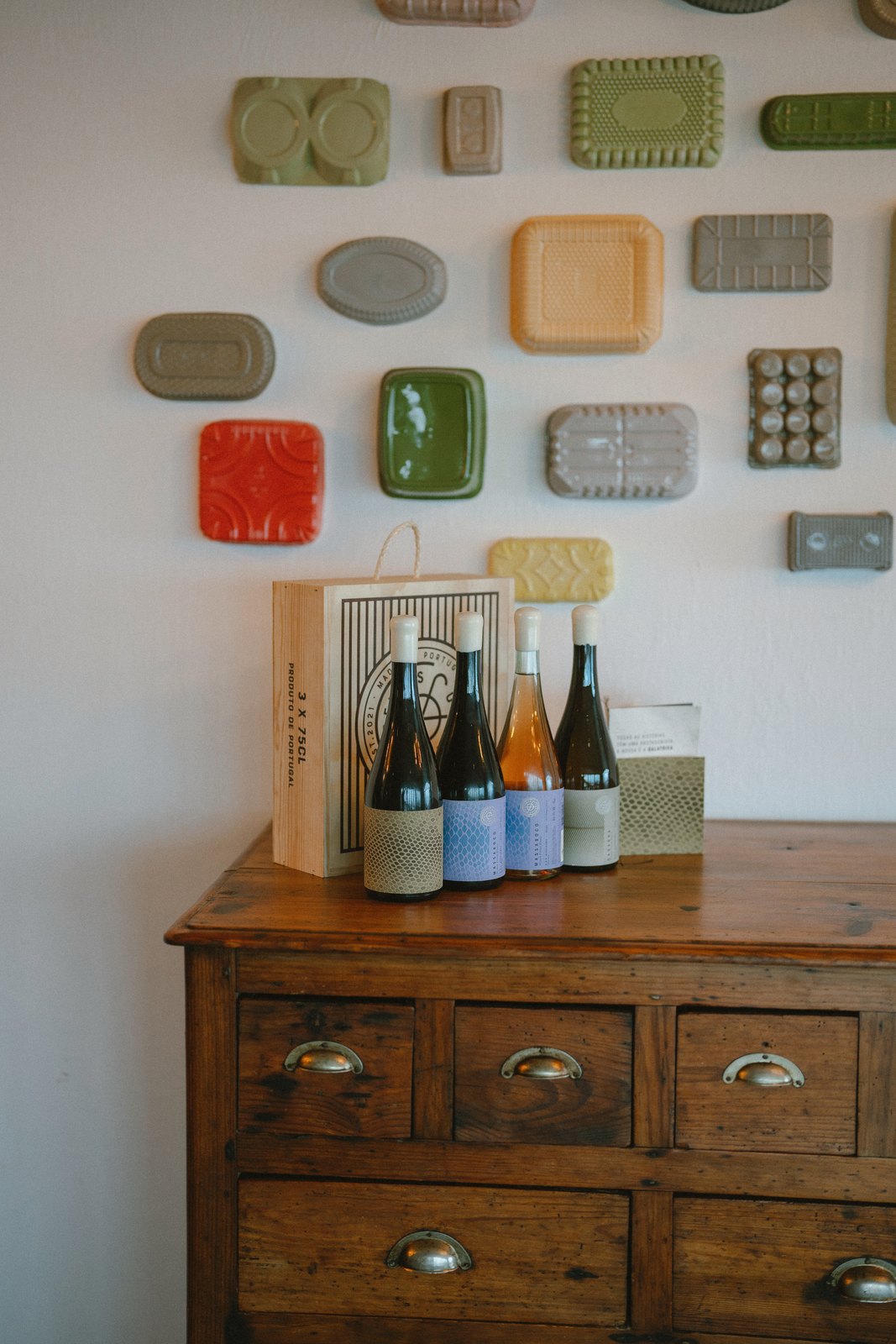 Socalco Nature Calheta — Ceramic plates on a white wall above an antique chest of drawers with wine bottles