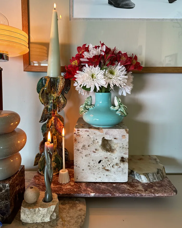 A travertine block holding a pale blue ceramic vase of white and red flowers, beside lit candles in a leaf-shaped holder, on a marble tray