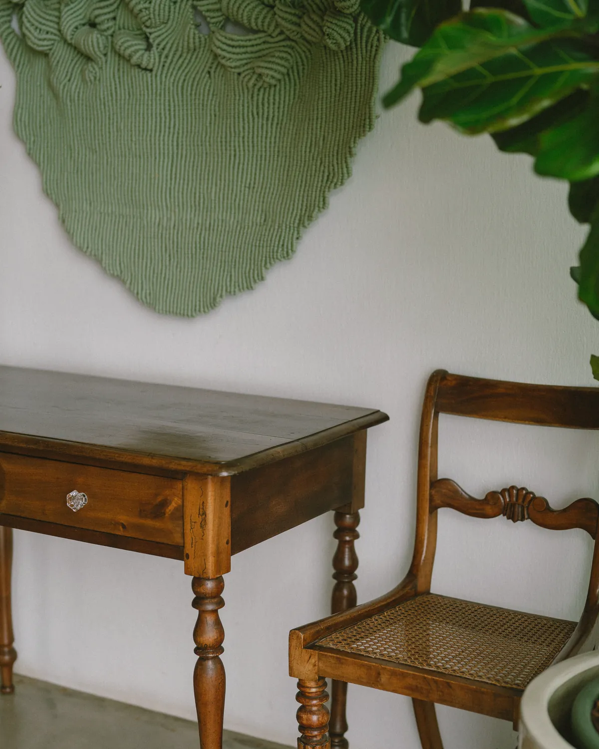 A sculpted green textile hanging on a white wall above an antique wooden desk and a cane-seat chair, at Socalco Nature Calheta